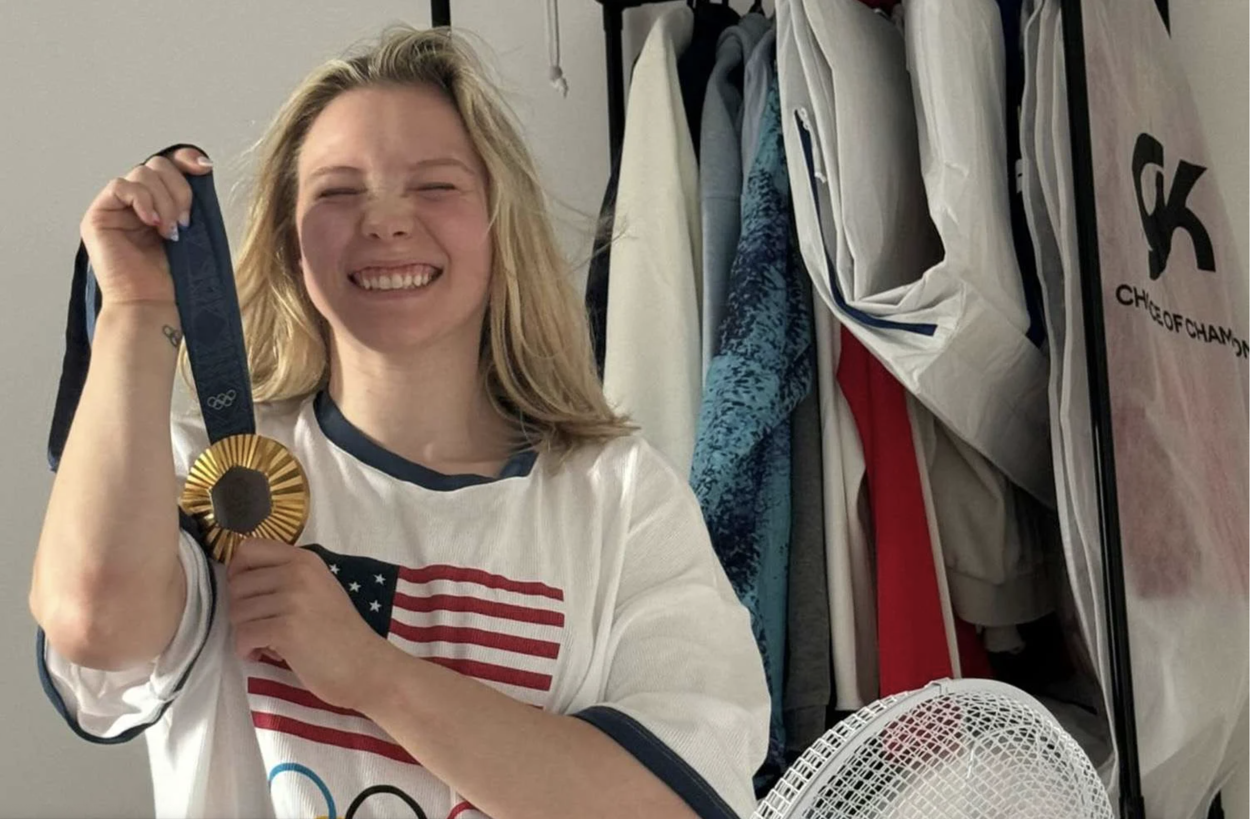 Jade Carey, a young female gymnast, is wearing the SKIMS Olympic oversized t-shirt pajama dress and holding up her new medal. Carey stands in front of a fan in the image, and looks fresh out of bed.